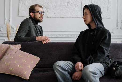 Jongen met capuchon zit op de bank en man met bril zit achter de bank. Ze kijken elkaar aan
