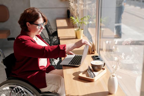 Vrouw met rood shirt in rolstoel zit aan tafel op haar laptop